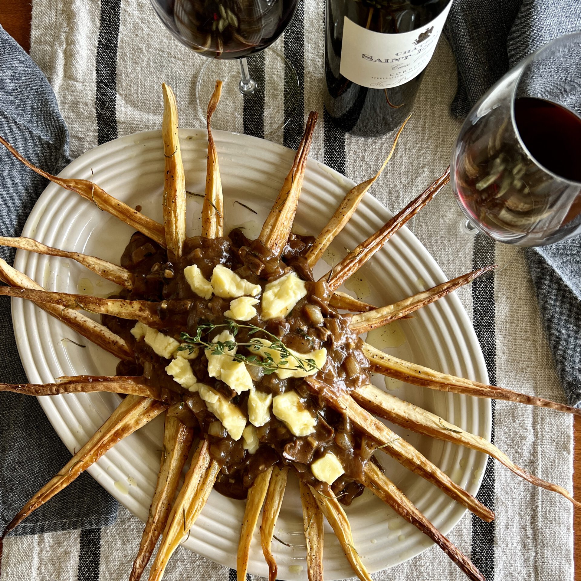 Parsnip Poutine with Rich Mushroom Gravy on a platter, surrounded by wine bottle and glasses.
