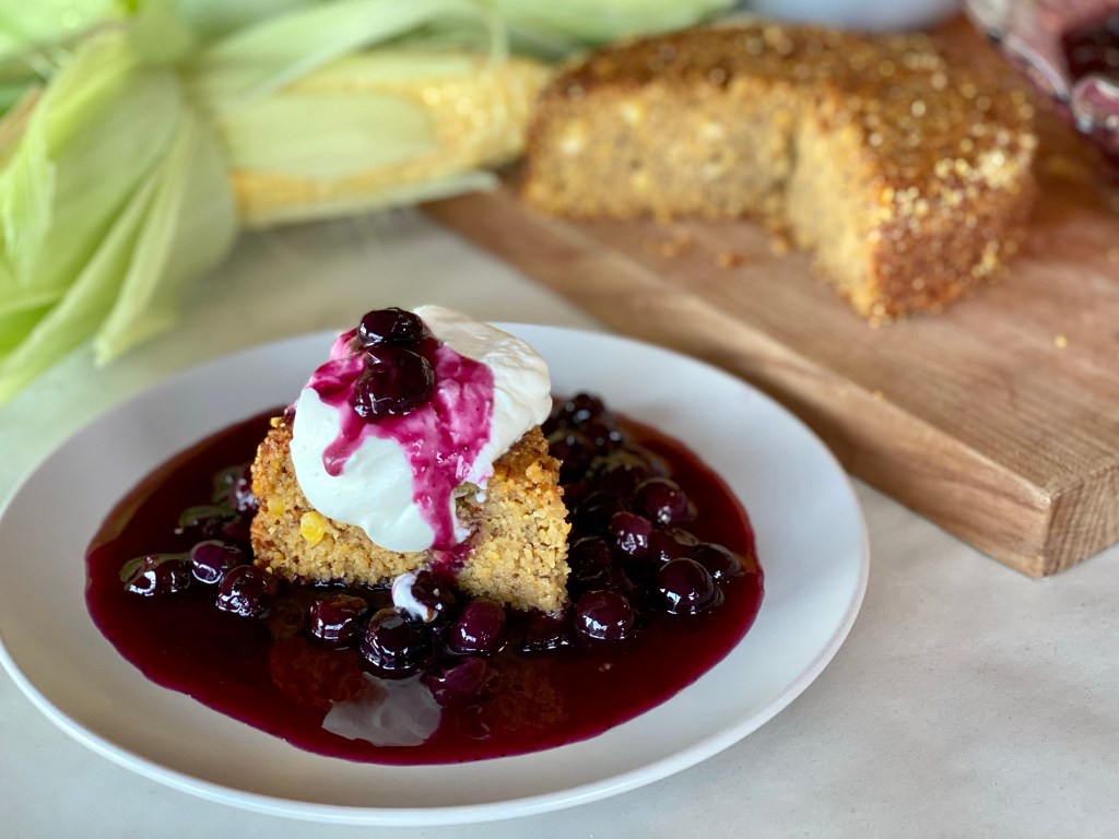 A slice of Sweet Corn Buttermilk Cake with blueberry compote and whipped cream on a plate.
